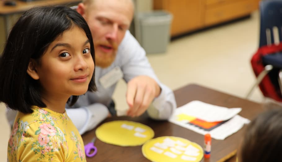 A student smiles directly at the viewer. A helpful volunteer is in the background